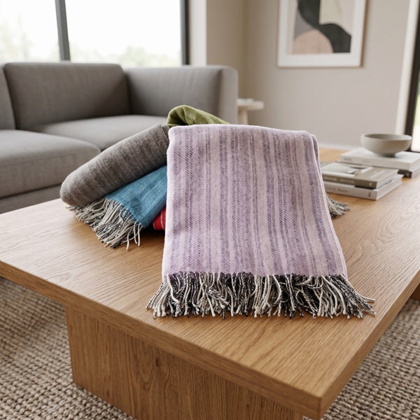 Indulge in the cosy comfort and timeless beauty of the Frenni Welsh Blanket bringing a touch of understated luxury and exceptional craftsmanship into your home. A folded blush pink throw blanket with grey fringe is draped over a stack of other folded blankets on a wooden coffee table in a modern living room setting.