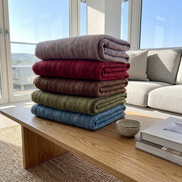 Welsh blankets and throws - Frenni. Five rolled knit throw blankets in pastel and earthy colours are arranged on a wooden coffee table in a modern living room setting. A stack of five folded textured blankets in muted colours (blush pink, carmine, conker, olive green, and aqua blue) arranged on a wooden coffee table in a bright, modern living room.