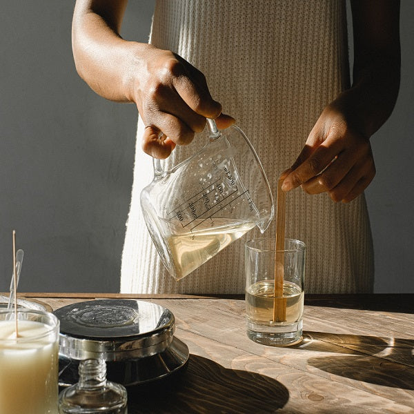 Candle making works - Person pouring liquid from a glass container into a glass on a wooden table with a candle and coaster.