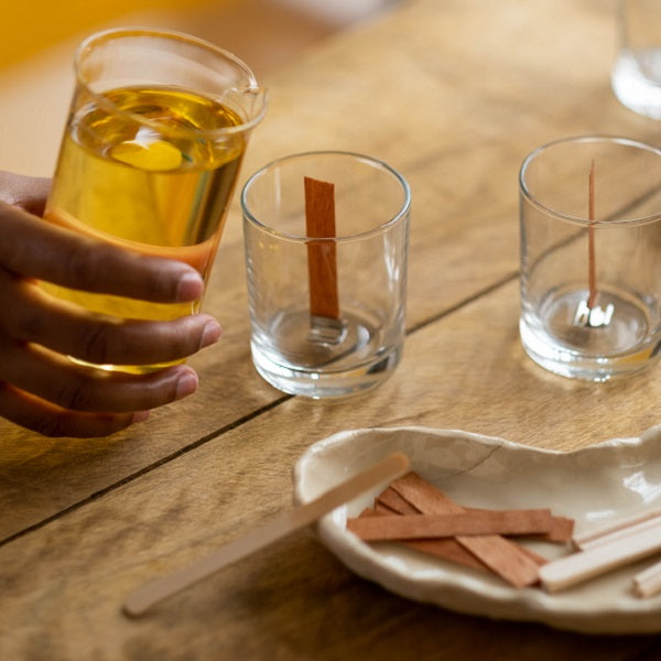 Candle making workshops - Hand holding a glass of amber liquid with wicks on a wooden table.