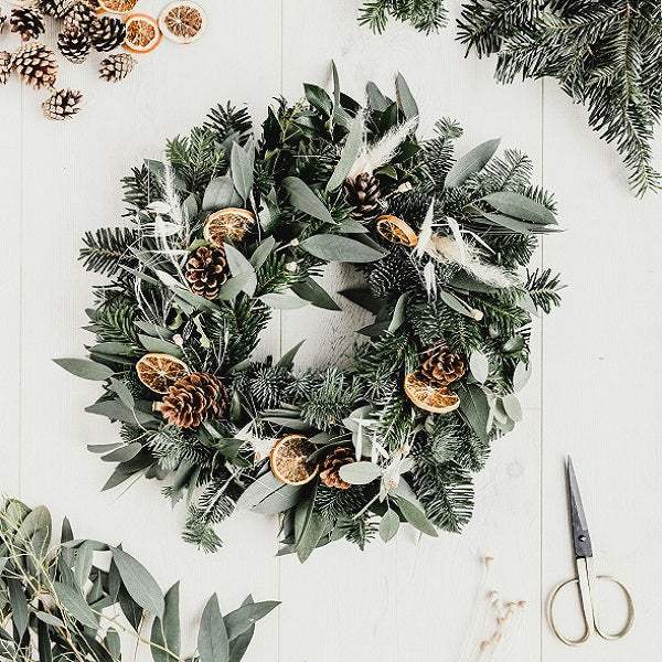 Cardiff Workshops. Christmas Wreath Making Workshop. Decorative wreath with greenery, pinecones, and dried oranges on a white background.