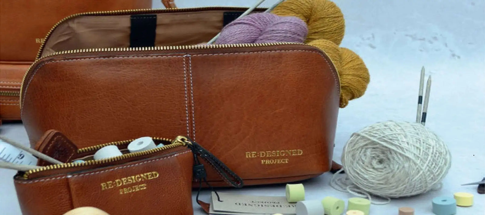 A brown leather zippered pouch with gold zipper and 'RE DESIGNED PROJECT' branding, shown alongside a matching small pouch, knitting needles, and balls of yarn on a white surface.
