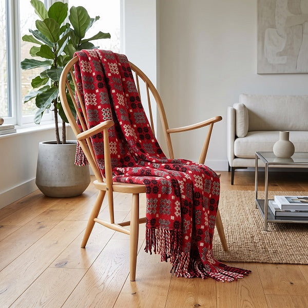 Welsh Blanket - Draig Goch - Red Dragon. A red patterned throw blanket with fringe is draped over a light wood armchair in a bright, modern living room with a potted fiddle-leaf fig, a beige sofa, and a glass coffee table.