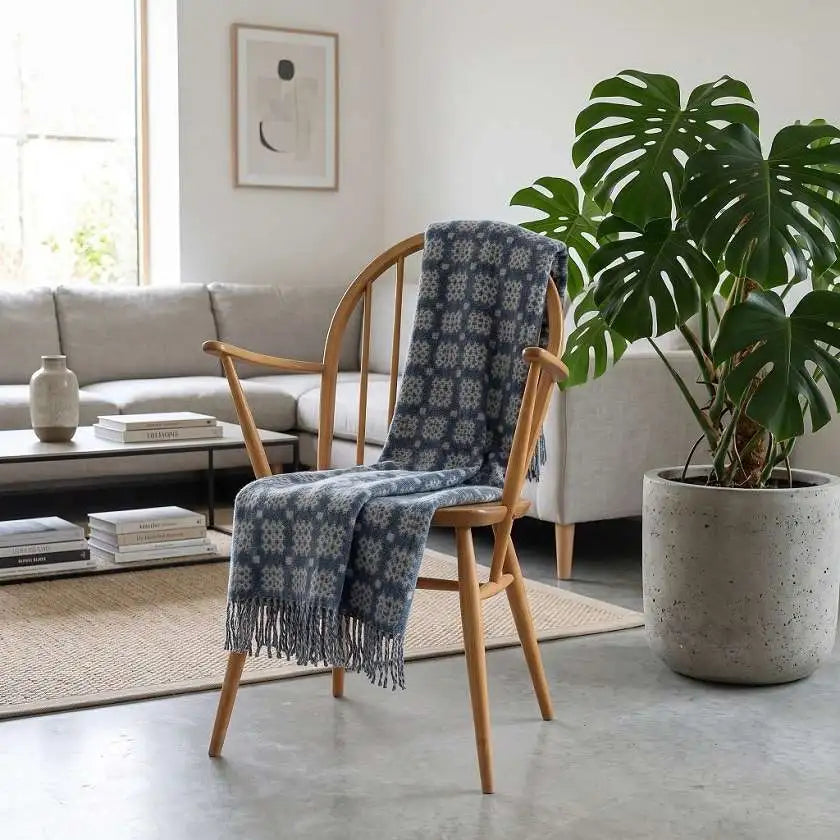 Welsh blankets named Craig Goch. A blue and white patterned fringed throw blanket is draped over a light wood armchair in a bright, modern living room setting next to a large potted monstera plant.