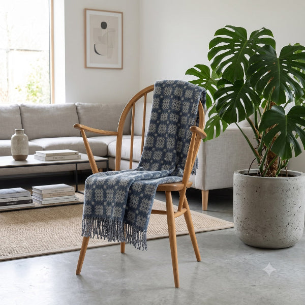 Welsh blanket named Craig Goch. A denim blue and white patterned fringed throw blanket is draped over a wooden armchair in a bright, modern living room with a grey sofa and a large green potted plant.